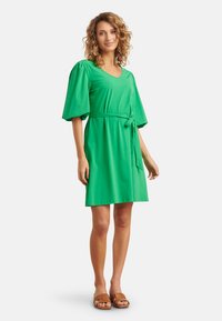 Groene jurk met korte mouwen van zachte stof. Heeft een v-hals en een gebonden taille, met een relaxed fit en uitlopende mouwen. Platte sandalen zichtbaar.