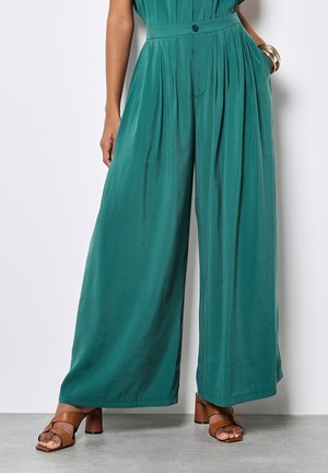 Szafirowe, wysokie w talii spodnie z szerokimi nogawkami i plisami, zestawione z brązowymi sandałami na obcasie oraz złotymi bransoletkami na prawej ręce.