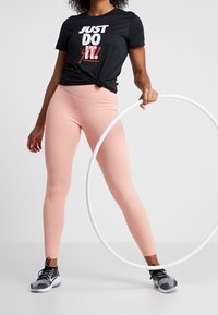 Svart T-shirt med texten "JUST DO IT!", kombinerad med ljusrosa leggings. Modellen håller en vit rockring och har på sig svarta och grå sportskor.