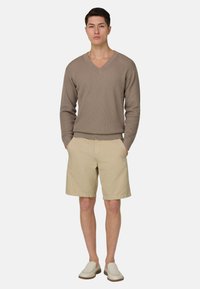 Homme portant un pull en V taupe, un short beige mi-cuisses et des chaussures blanches sans lacets, debout les mains dans les poches sur fond blanc.