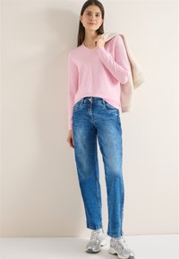 Femme portant un pull à manches longues rose clair, un jean bleu, des baskets argentées, tenant une veste beige sur l'épaule devant un fond uni.