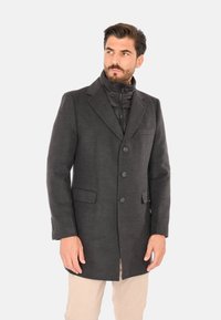 Uomo che indossa un soprabito grigio scuro abbottonato con colletto trapuntato nero con cerniera e pantaloni beige, che guarda verso sinistra.