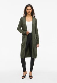Long cardigan vert olive fabriqué en matériau doux, avec une ouverture à l'avant, deux grandes poches et des poignets côtelés. Porté sur un haut blanc.