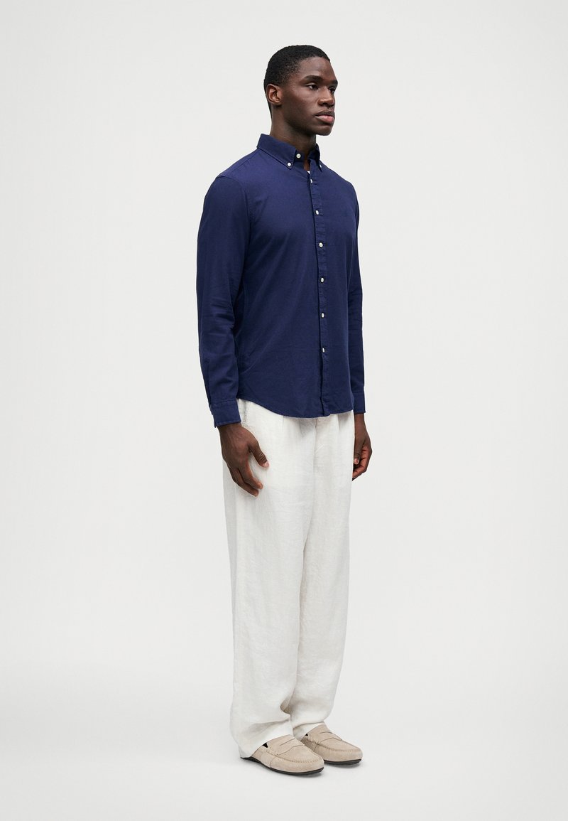 Mørkeblå langærmet skjorte med knapper og krave, parret med løse kremfarvede linnedbukser og lyse slip-on sko. Enkel, afslappet pasform.