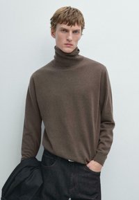 Brązowy sweter z golfem z miękkiej tkaniny, z długimi rękawami i dopasowanym krojem. Nosi się go z ciemnymi dżinsami i czarną kurtką, co podkreśla minimalistyczny styl.
