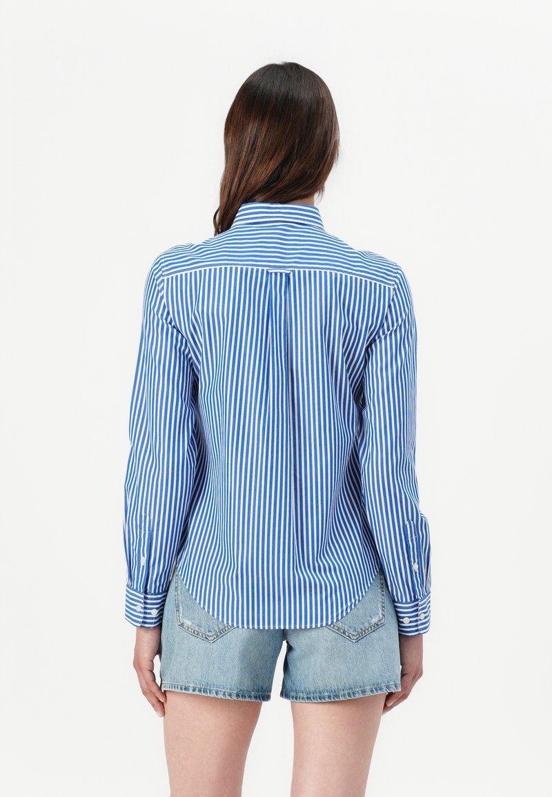 Rückansicht eines blau-weiß gestreiften Hemdes mit langen Ärmeln, das einen abgerundeten Saum und einen maßgeschneiderten Schnitt aufweist, kombiniert mit Jeans-Shorts.