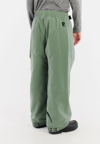 Groene waterdichte broek met een elastische tailleband, losse pasvorm, drukknoopsluitingen bij de enkels en een logodetail in de buurt van de achterzak.