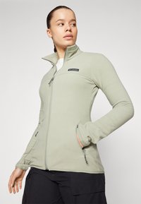 Vaaleanvihreä fleece-takki, jossa on korkea kaulus, vetoketjukiinnitys ja sivutaskut. Takissa on logo-patch rintataskussa ja joustavat hihansuut.