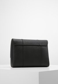 Clutch bag in pelle nera texture con chiusura a pattina, mostrato in posizione verticale su un pedestal bianco su uno sfondo semplice.