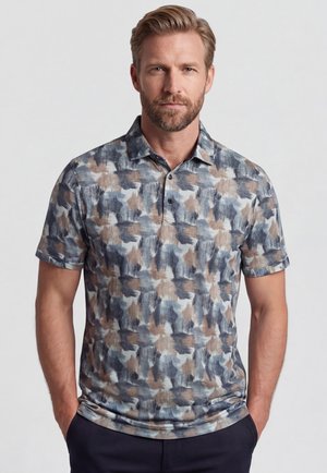 Man met baard die een kort mouwhemd met kraag draagt met een abstract patroon van blauwe, beige en grijze penseelstreken, handen in de zakken, neutrale achtergrond.