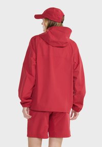 Rode waterdichte jas met een capuchon, lange mouwen en elastische manchetten, gecombineerd met bijpassende shorts en een pet, allemaal gemaakt van glad, lichtgewicht materiaal.