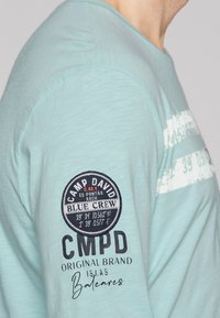 Licht turquoise langemouwen shirt met een ronde patch met "Camp David" en "Blue Crew", plus een geprinte ontwerp met strepen en tekst.