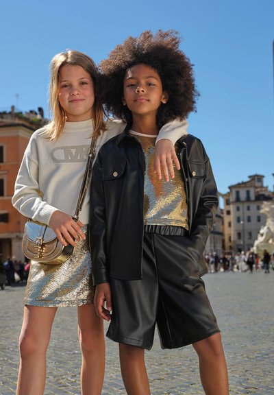 Due ragazze sicure di sé stanno all'aperto in una piazza urbana soleggiata, una con un braccio attorno all'altra, vestite con eleganti outfit oro e nero.