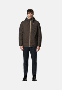 Giacca imbottita marrone con cappuccio, cerniera gialla e bianca, con logo sul petto a sinistra. Indossata con pantaloni eleganti blu navy e scarpe nere.