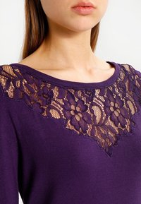 Maglia a maniche lunghe viola con un bordo in pizzo floreale sul colletto, con una finitura in tessuto liscio e una vestibilità comoda.