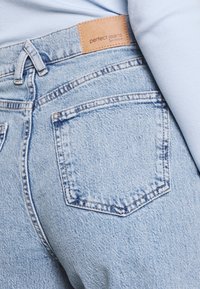 Světle modré džíny z denimu s texturovaným povrchem, s koženou nášivkou označenou "dokonalé džíny" a dvěma zadními kapsami se švy.