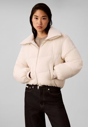 Kvinde med mørkt hår iført en cremefarvet dunjakke med hænderne i lommerne og sorte højtaljede jeans mod en lysegrå baggrund.