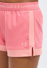 Κοντινό πλάνο ροζ αθλητικών σορτς Under Armour με ελαστική μέση, πλαϊνή τσέπη, λογότυπο και μικρή πλαϊνή σχισμή στον μηρό ενός ατόμου.