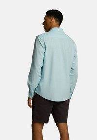 Jasnoniebieska koszula z długim rękawem, z kołnierzykiem na guziki i regulowanymi mankietami, noszona z ciemnymi szortami. Gładka faktura, casualowy krój, widok z tyłu.