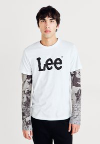 Hvit bomull t-skjorte med en stor svart "Lee" logo. Kombinert med grå langermede skjorter med mørke mønstre og teksturer. Svarte bukser.