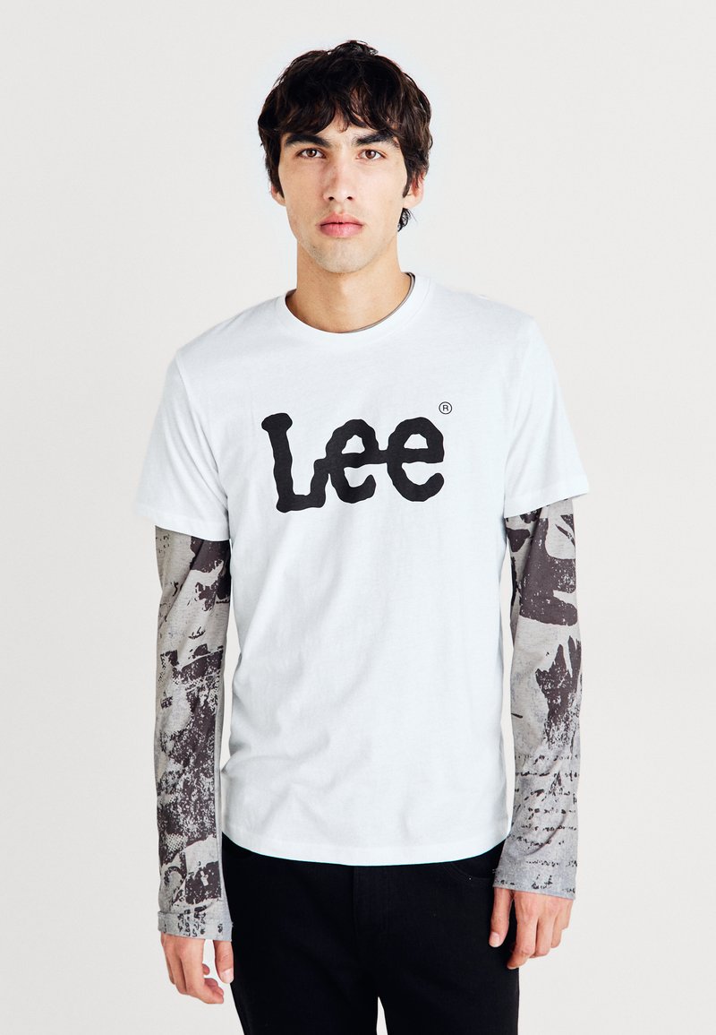 Hvit bomull t-skjorte med en stor svart "Lee" logo. Kombinert med grå langermede skjorter med mørke mønstre og teksturer. Svarte bukser.