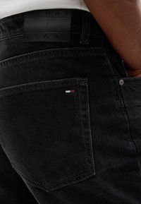 Κοντινό πλάνο σε μαύρο τζιν Tommy Jeans με πίσω τσέπη και μικρό κόκκινο, άσπρο και μπλε λογότυπο στην τσέπη.