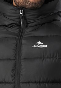 Veste matelassée noire avec fermeture éclair sur le devant, arborant un logo blanc "Helvetica Designed in the Alps" et un graphisme de montagne sur la poitrine.