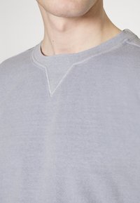 Vaaleanharmaa collegepaita V-mallisella pääntiellä, valmistettu pehmeästä puuvillasekoitteesta, jossa on hillittyjä ompeluaiheita kauluksen kohdalla.