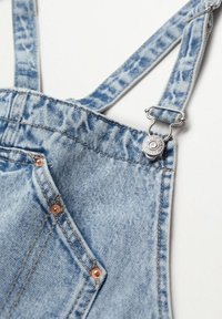 Bretelle de salopette en denim bleu clair avec boucle métallique et bouton-pression, présentant des coutures visibles et des rivets en cuivre sur le coin de la poche.