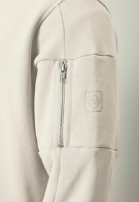 Sudadera de color beige claro con un bolsillo con cremallera en la manga, textura acolchada y un sutil parche con logo en relieve.