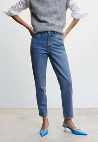 Szary sweter dzianinowy, biała koszula, niebieskie spodnie z wysokim stanem oraz niebieskie szpilki z wąskim noskiem wyeksponowane na neutralnym tle.