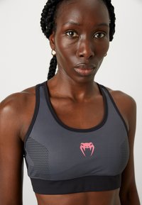 Sporta krūšturis tumši pelēkā krāsā ar rozā kobras logo. Iespējas ietver acs paneļus sānos, gludu tekstūru un pieguļošu dizainu, lai nodrošinātu atbalstu.