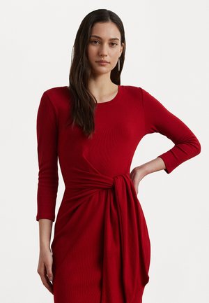 Red knit dress with long sleeves, a round neckline, and a twisted knot detail at the waist, showcasing a smooth texture and flowing fabric.