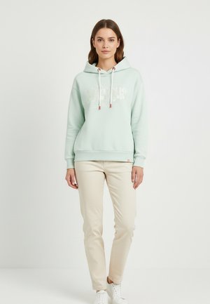 Frau steht und trägt einen hellgrünen Hoodie mit weißen Kordeln, beige Hose und weiße Turnschuhe vor einem schlichten hellen Hintergrund.