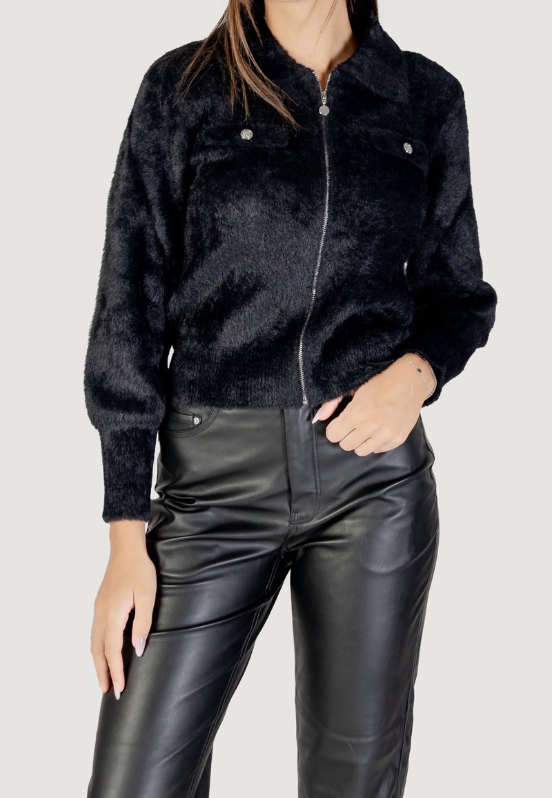 Femme portant une veste noire à fermeture éclair en tissu duveteux avec des poches poitrine boutonnées et un pantalon noir taille haute en cuir synthétique.