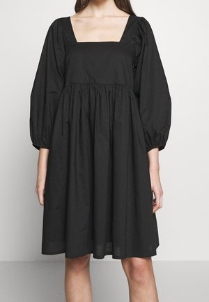 Femme portant une robe noire mi-longue avec des manches bouffantes trois-quarts et un décolleté carré, debout devant un fond uni.