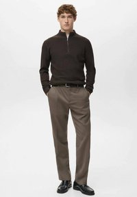 Suéter marrón acanalado con cremallera en el cuello combinado con pantalones plisados de tono marrón claro; el modelo lleva zapatos negros; fondo minimalista.