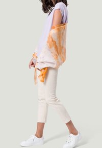 Blouse lilas clair associée à une écharpe longue avec des accents orange, un pantalon court crème et des baskets blanches, présentant des textures douces et un tissu léger.