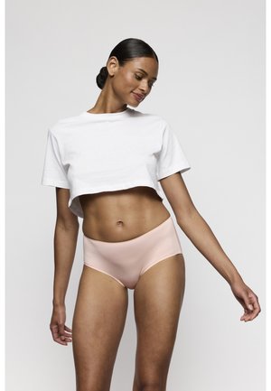 Vrouw met donker haar in een knot, draagt een wit cropped t-shirt en lichtroze naadloos ondergoed, staand tegen een effen grijze achtergrond.