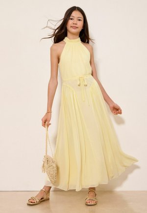 Mladá žena nosí bezrukávové bledozlaté maxi šaty s halter krkom, drží pletenú tašku, stojí pred neutrálnym pozadím s sandálmi.