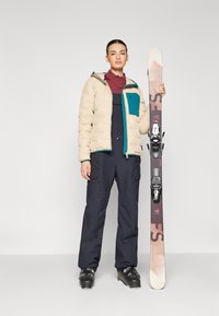 Puffet beige jakke med teal-lomme, maroon topp, marineblå snøbukser og svarte skistøvler. Holder en multikolor skistav med bindinger.