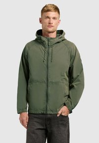 Homme portant une veste zippée à capuche vert olive et un jean foncé, debout avec une main dans la poche, sur un fond uni.