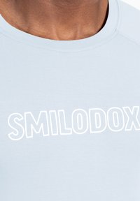 Camiseta de color azul claro con cuello redondo, que presenta un logotipo en texto blanco "SMILODOX" de forma destacada en el pecho. Tela suave y lisa.