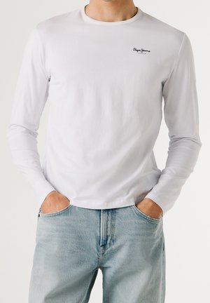 Homme portant un t-shirt blanc à manches longues Pepe Jeans et un jean bleu clair, mains dans les poches, devant un fond uni.