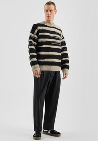 Zwart-beige gestreepte trui met een geribbelde halslijn en manchetten, gecombineerd met losse zwarte broek en zwarte sneakers.