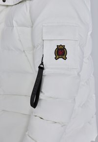 Valge puff jakk, millel on lukuga sääre tasku, millel on broderitud logo, ja must brändi silt, mis on kinnitatud luku tõmbesse.
