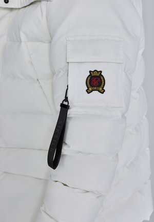 Bílá péřová bunda s kapsou na zip na rukávu, vyšívaným logem a černým štítkem značky připevněným na táhlu zipu.