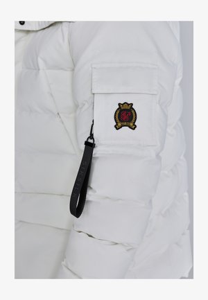 Valge puff jakk, millel on lukuga sääre tasku, millel on broderitud logo, ja must brändi silt, mis on kinnitatud luku tõmbesse.