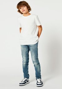 Balts kokvilnas T-krekls, zilas nodilušas šauras džinsa bikses un melni baltas sporta kurpes. Modelis stāv ar rokām kabatās, neitrāla fona priekšā.