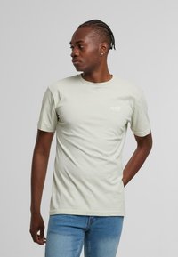 Hellgrünes Baumwoll-T-Shirt mit kurzen Ärmeln, rundem Halsausschnitt und kleinem weißen Logo auf der Brust. Getragen mit eng sitzenden blauen Jeans.